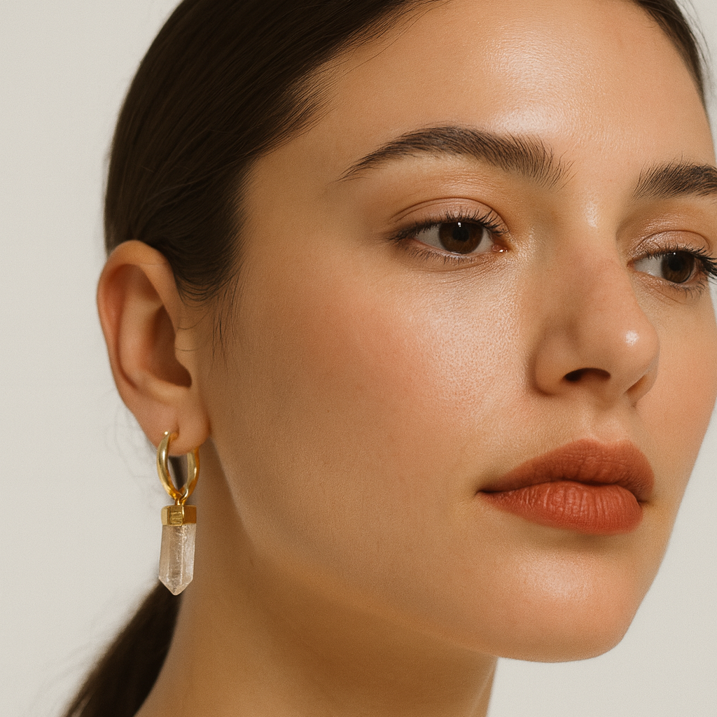 Close-up of a woman wearing gold earrings with a clear stone on a neutral background