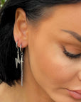 A close-up image of a woman's ear wearing a silver dangle earring with a cross and lightening bolt design.