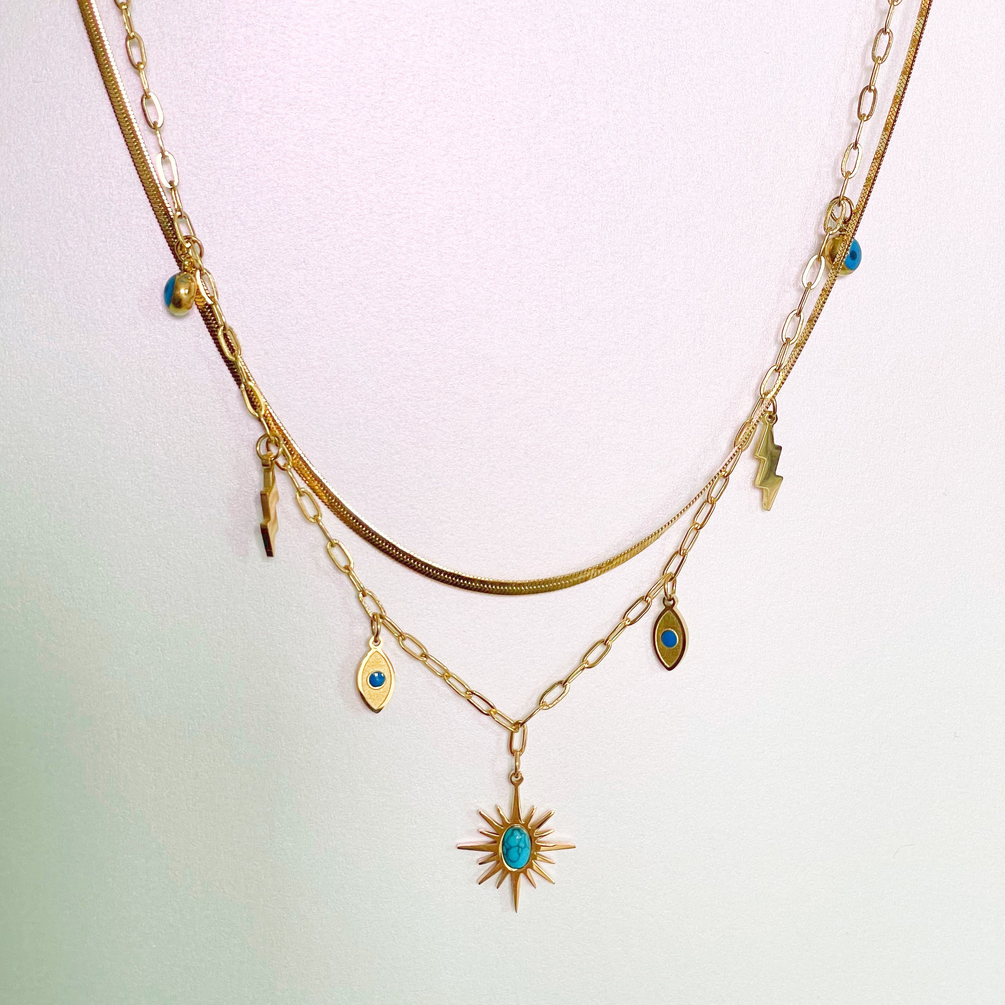 An 18k gold plated necklace with a sunburst pendant and layered chains, featuring small charms including an evil eye and lightning bolt, displayed against a light background.